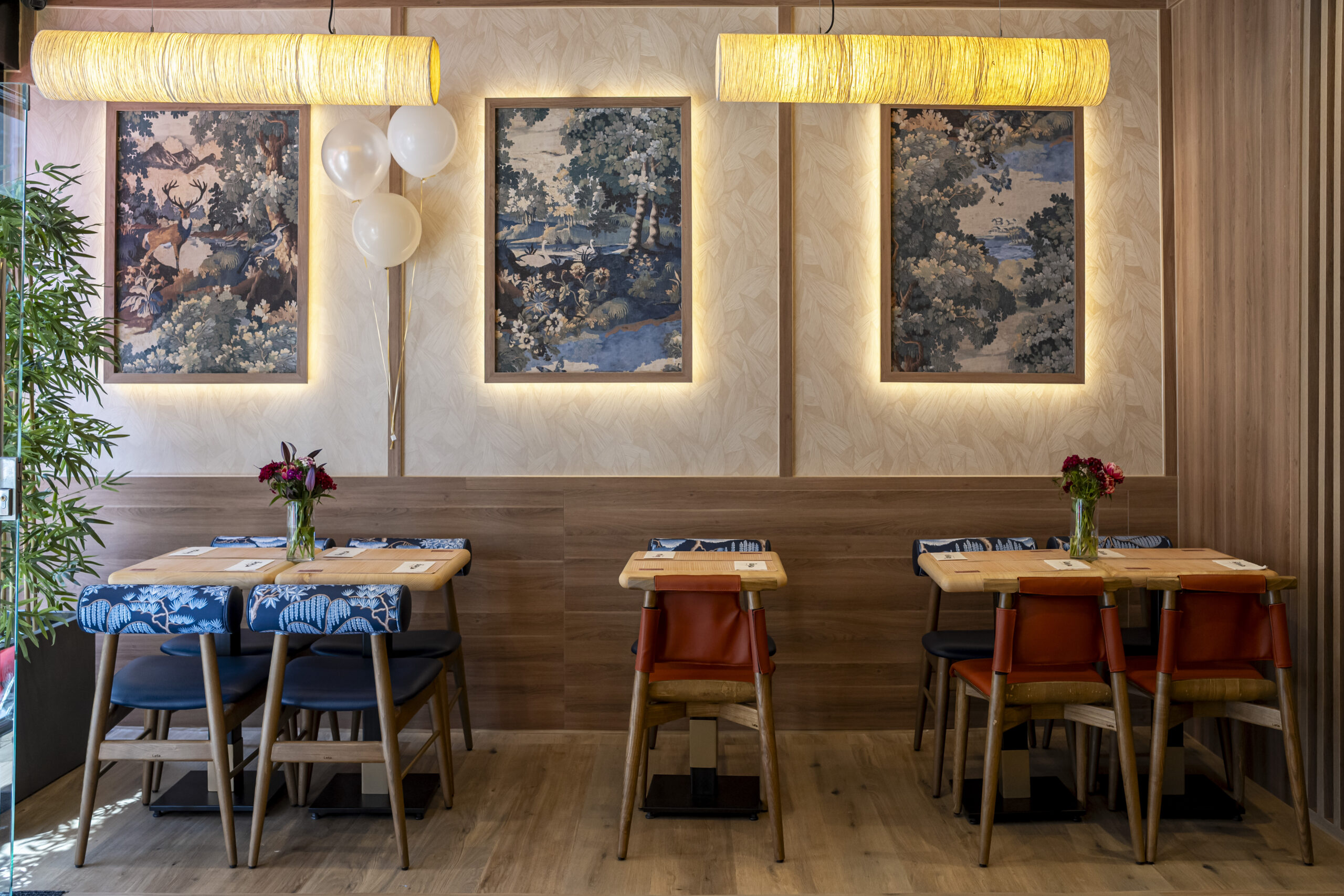 Cozy dining area with wooden tables and blue-patterned chairs, three framed nature tapestries on a beige paneled wall, warm hanging lights