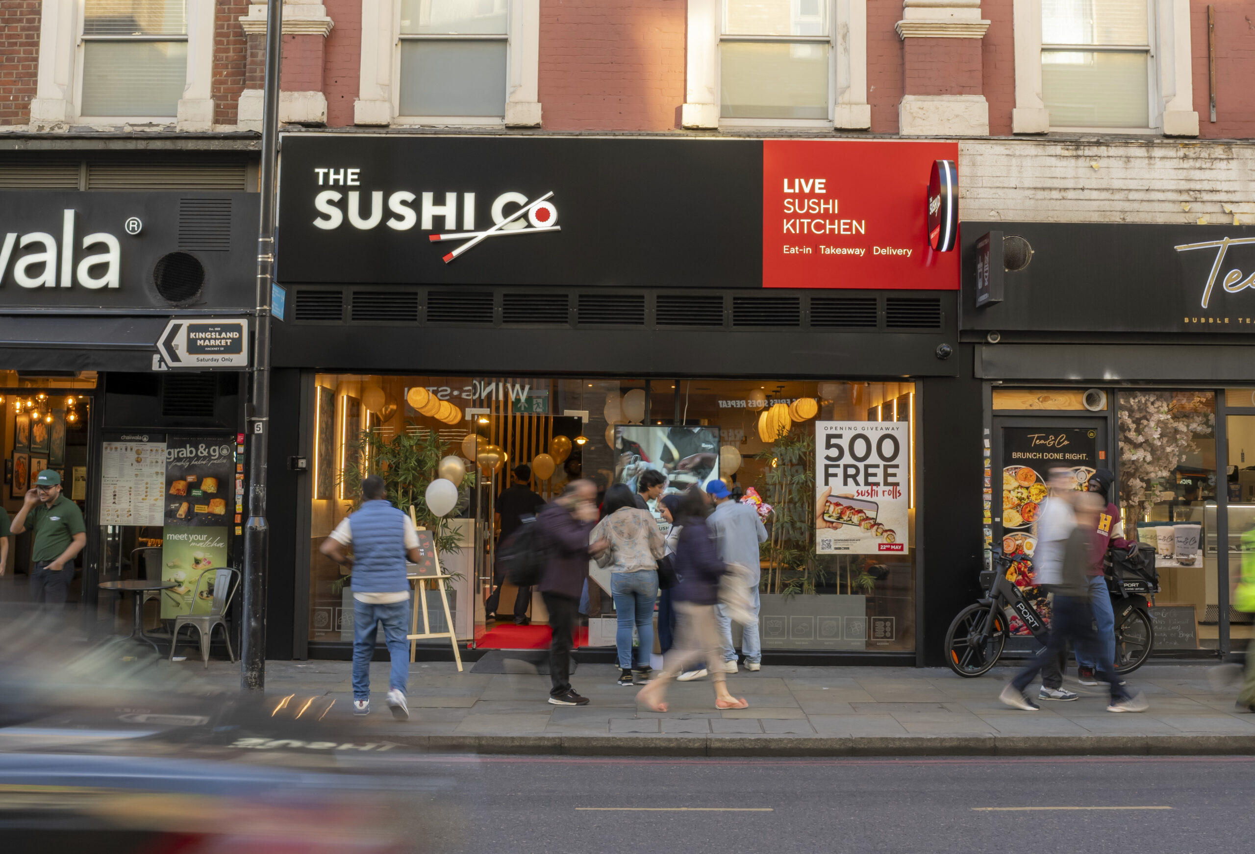 Street view of a row of shops with a red-brick building and a black-and-red sushi restaurant sign reading 'The Sushi Co' and 'Live Sushi Kitchen' as people walk by on the sidewalk.