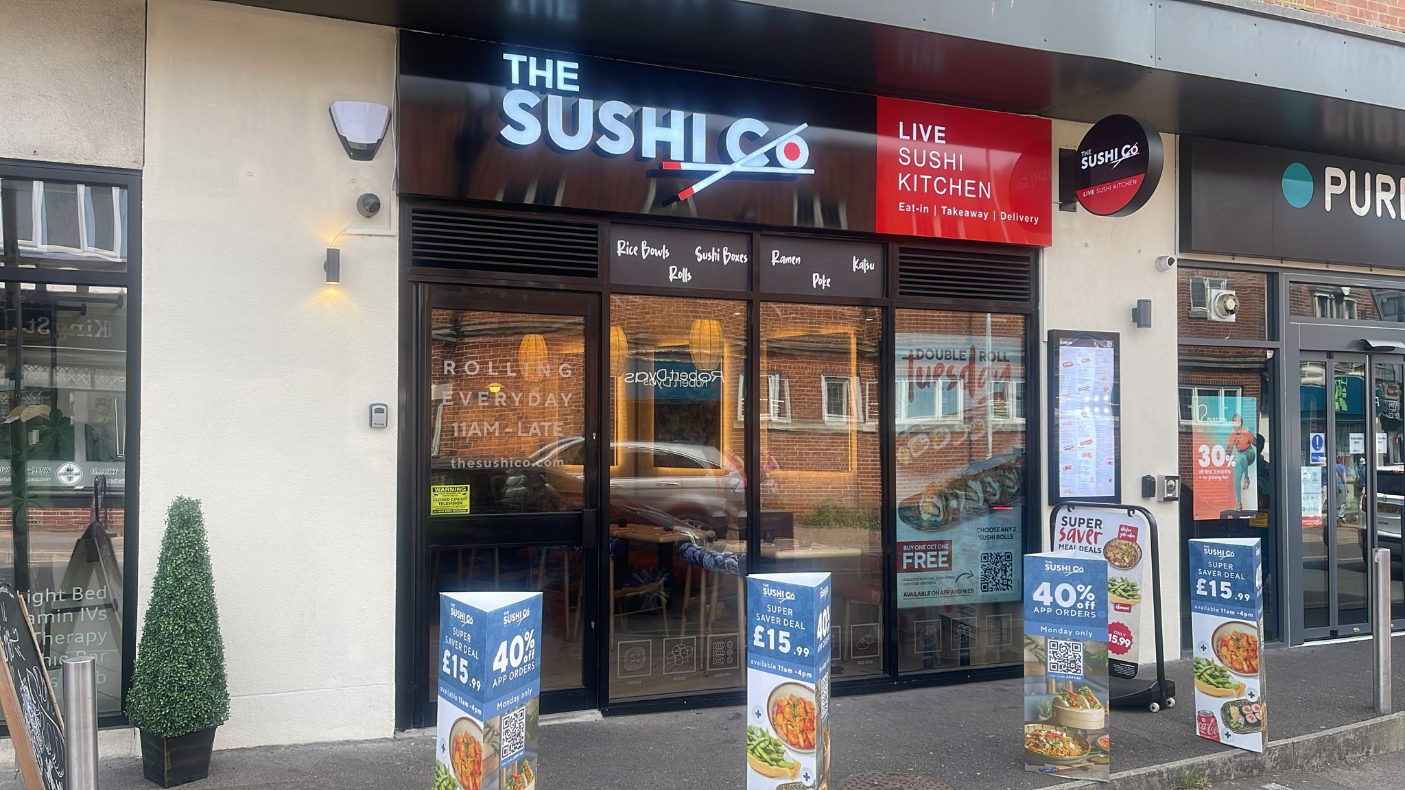 Front of a sushi restaurant with a large THE SUSHI sign and red Live Sushi Kitchen panel above the entrance; glass doors show interior seating and menu boards displaying items like rolls and ramen.
