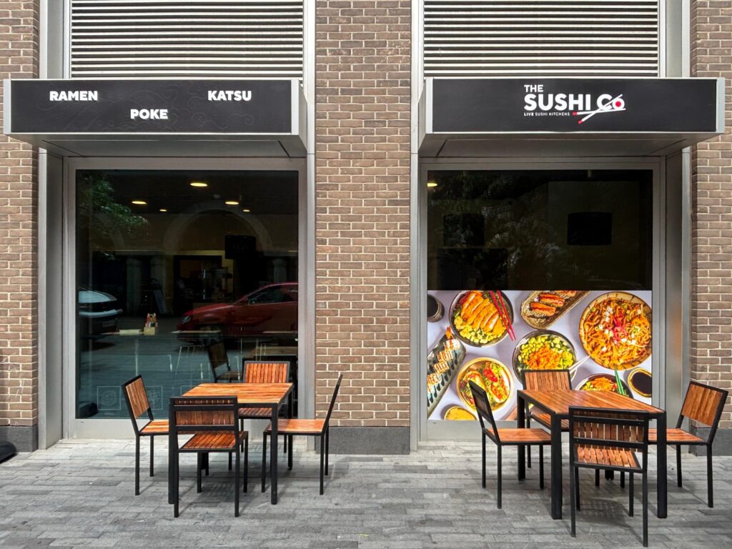 Restaurant storefronts with black signage and outdoor wooden tables; left sign reads ramen, poke, katsu, right sign advertises sushi, brick building façade.