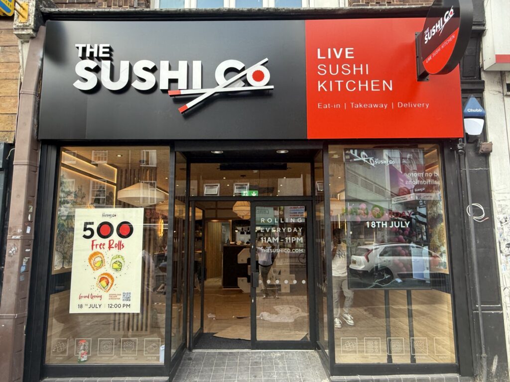 The Sushi Co storefront with black and red signage, glass doors, and posters advertising offers and the July 18 opening date.