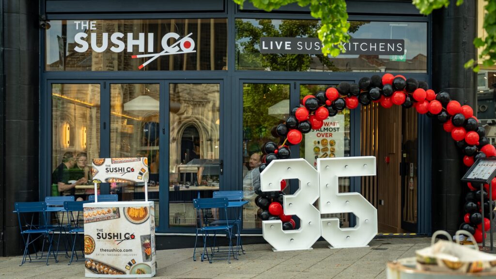 Exterior of The Sushi Co restaurant with a red-and-black balloon arch, blue outdoor tables and chairs outside the storefront signage visible above the door/ windows.