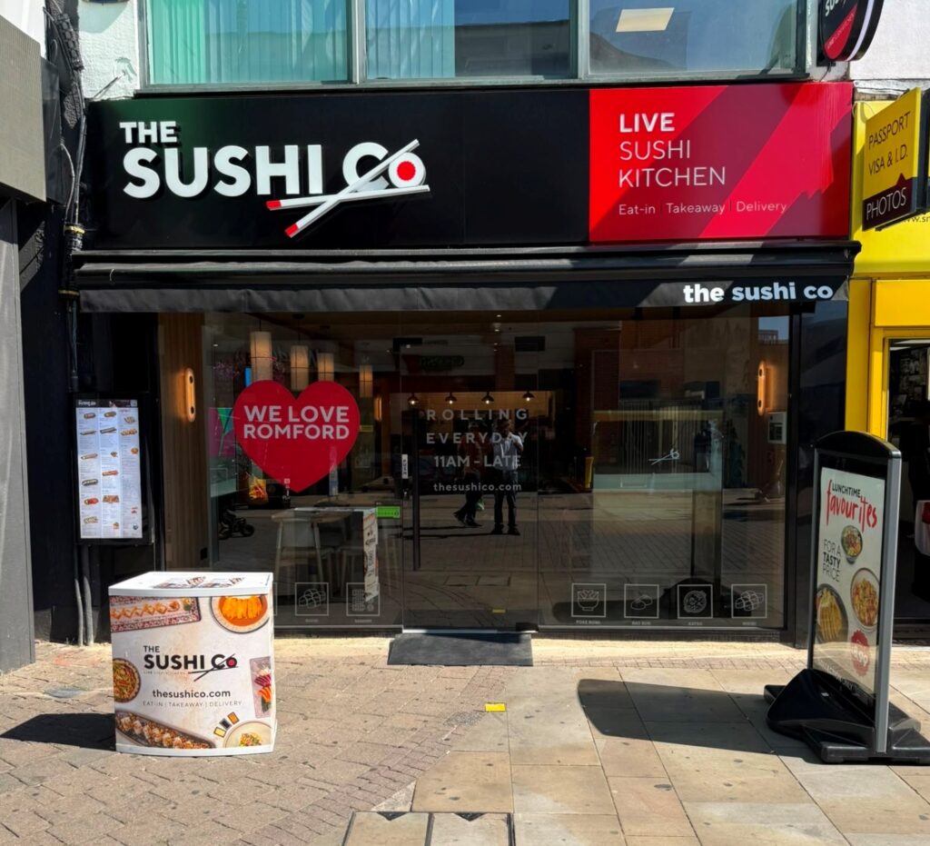 Front of The Sushi Co restaurant with black and red signage reading LIVE SUSHI KITCHEN; glass doors show a red heart graphic and interior seating inside Romford location.