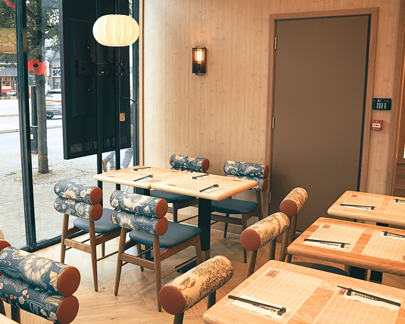 Cozy restaurant interior with wooden tables and blue floral-patterned chairs near a large window facing the street outside.