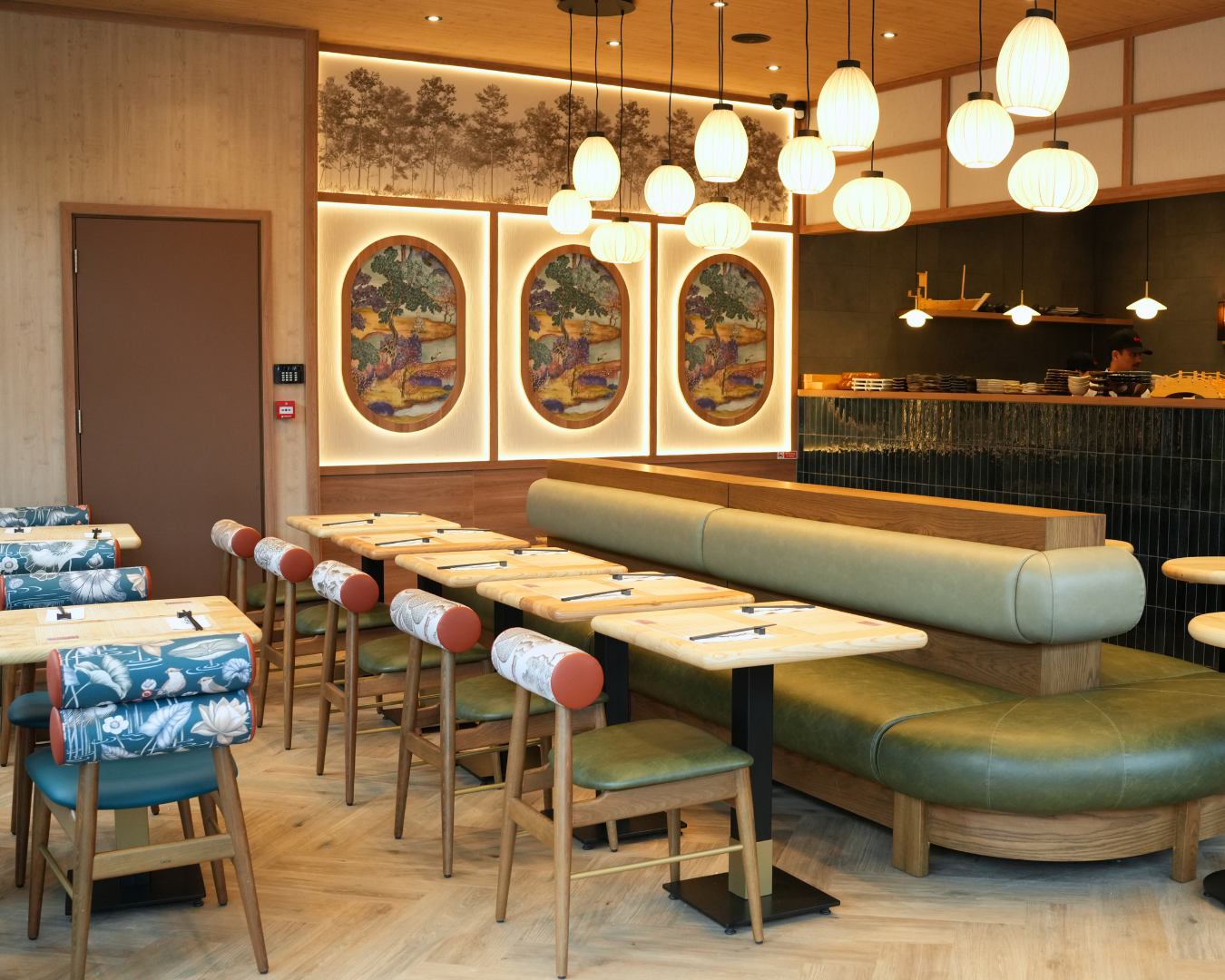 Modern dining area with light wood tables, patterned chairs, and a long green curved banquette under warm pendant lights.