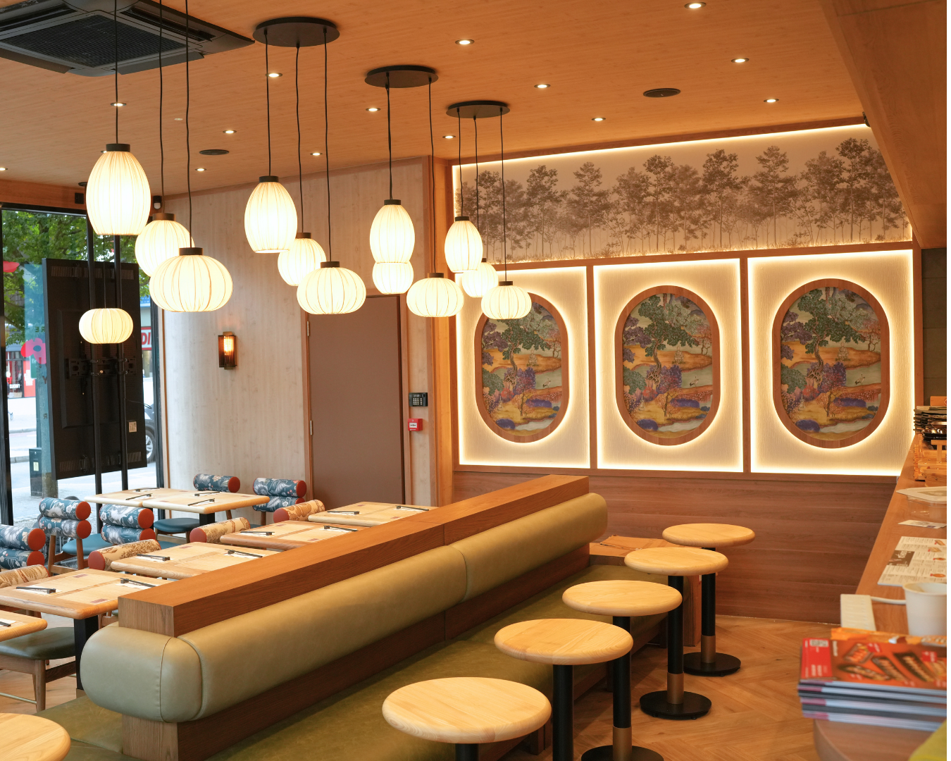 Warm, modern restaurant interior with multiple pendant lights, a long cushioned booth, and round stools along a wooden counter.