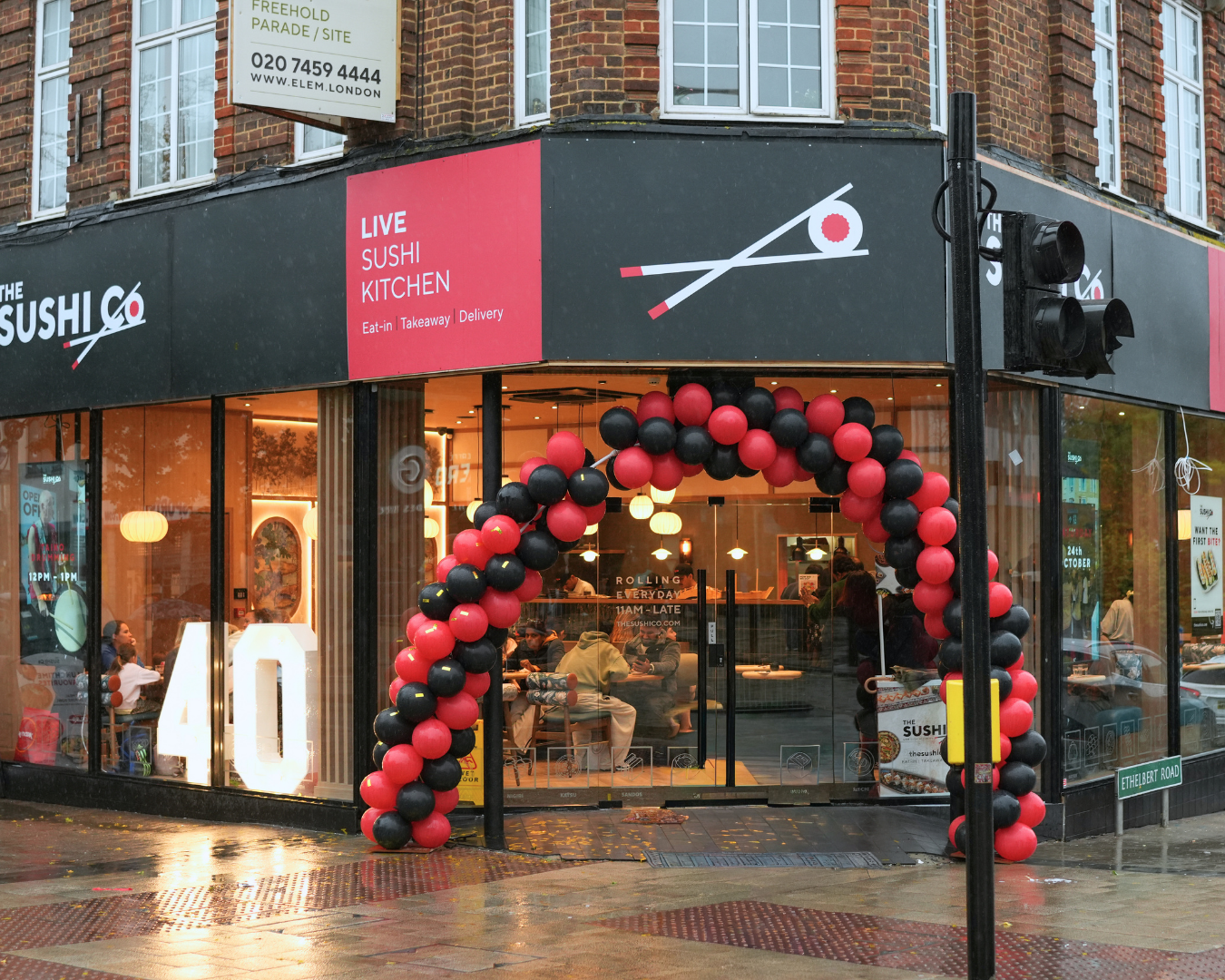 Sushi restaurant storefront with a pink sign 'LIVE SUSHI KITCHEN', a red/black balloon arch, and customers inside viewed through glass doors.