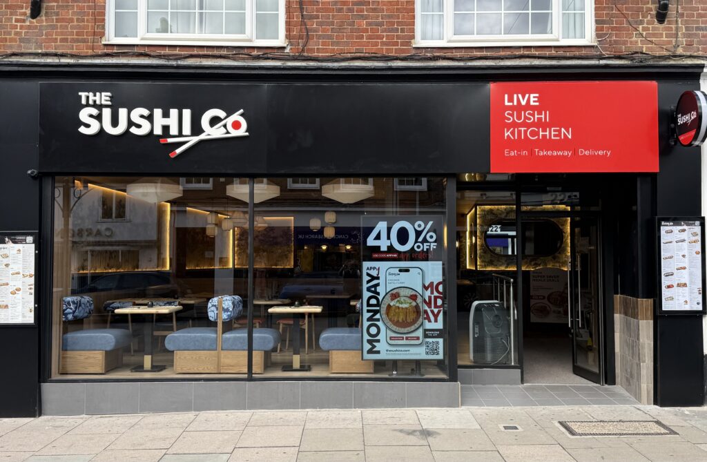 storefront of The Sushi Co restaurant with a black façade and a red Live Sushi Kitchen sign; dine-in seating visible through glass.