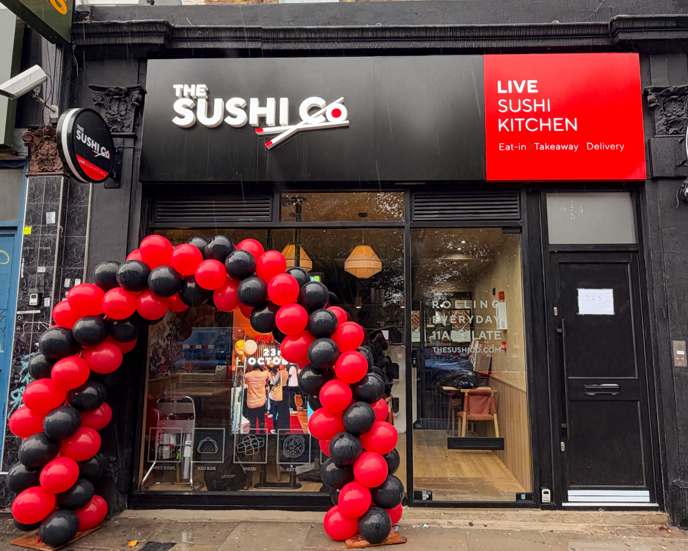 storefront for The Sushi Co with a black facade, bold white logo, and a red 'Live Sushi Kitchen' panel; red and black balloon arch at the entrance.