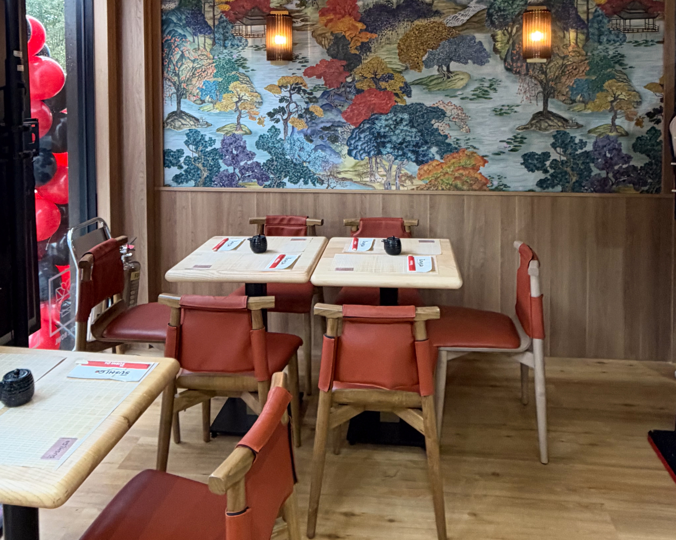 Cozy restaurant interior with light wood tables, red cushioned chairs, and a large colorful mural of trees on the back wall; warm hanging lanterns above.