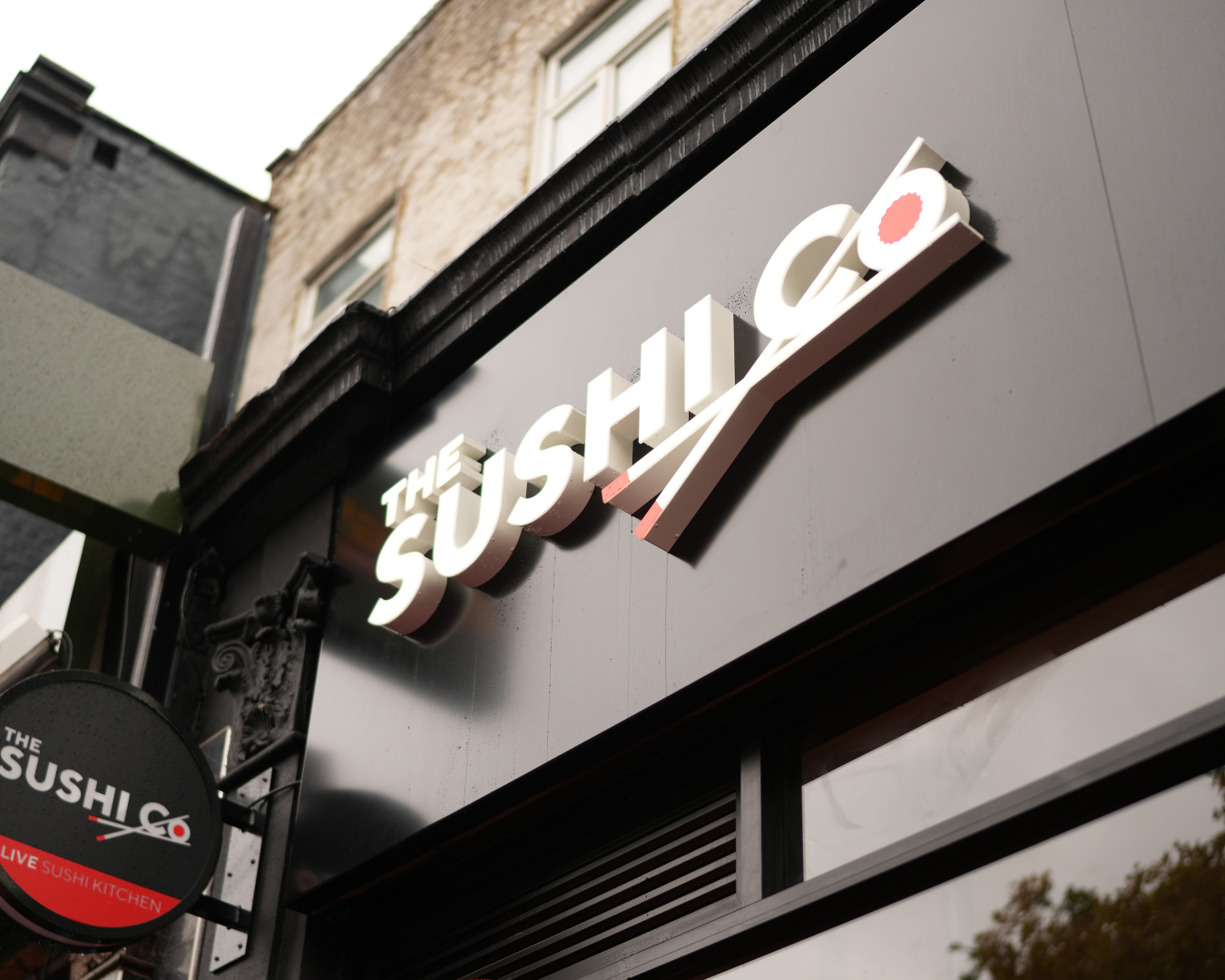 Exterior view of a sushi restaurant sign reading 'THE SUSHI CO' on a dark storefront panel.